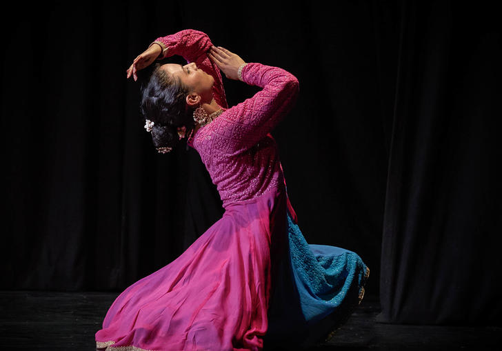 A dancer dressed in pink, kneels on a blacked out stage