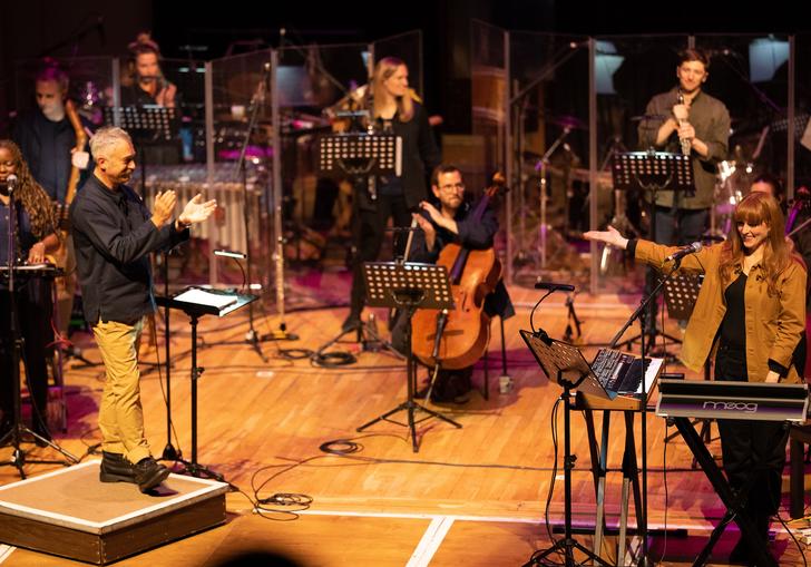 Hannah Peel performing with Paraorchestra. From behind a keyboard on the left of the stage she is gesturing to the ensemble who are applauding.