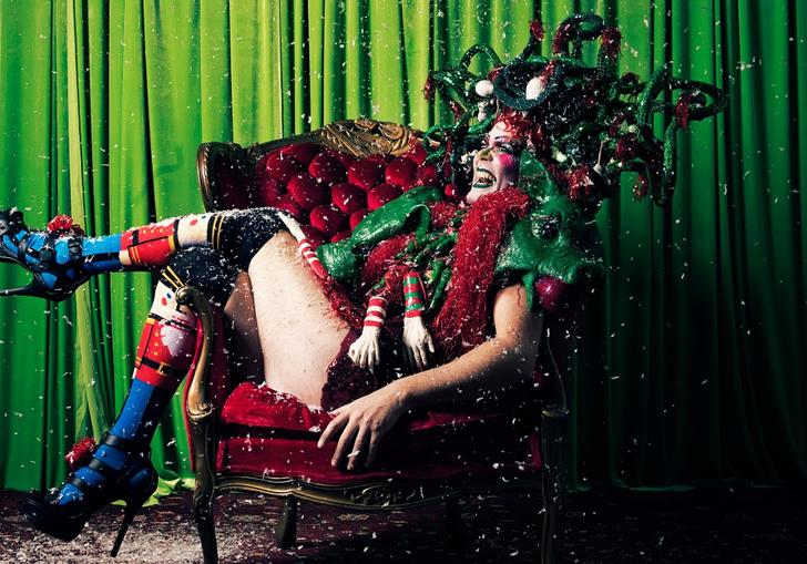 a colour photo of Taylor Mac in festive drag reclining over an armchair against a green backdrop