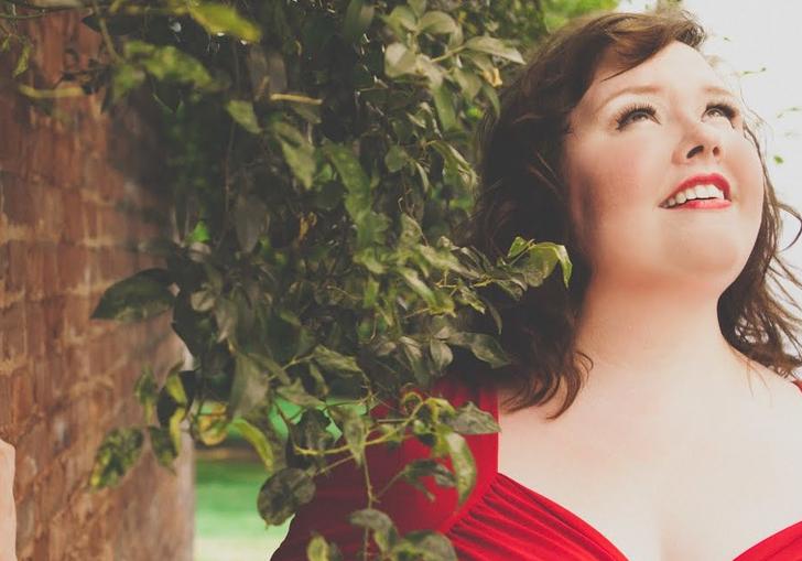Jamie Barton in a red dress beside a brick wall and a plant draping over her shoulder