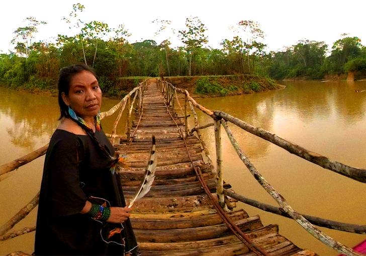 Hushuhu, the first woman shaman of the Yawanawá stands on a wooden bridge crossing a river The water is brown and she is holding a large white and black feather.