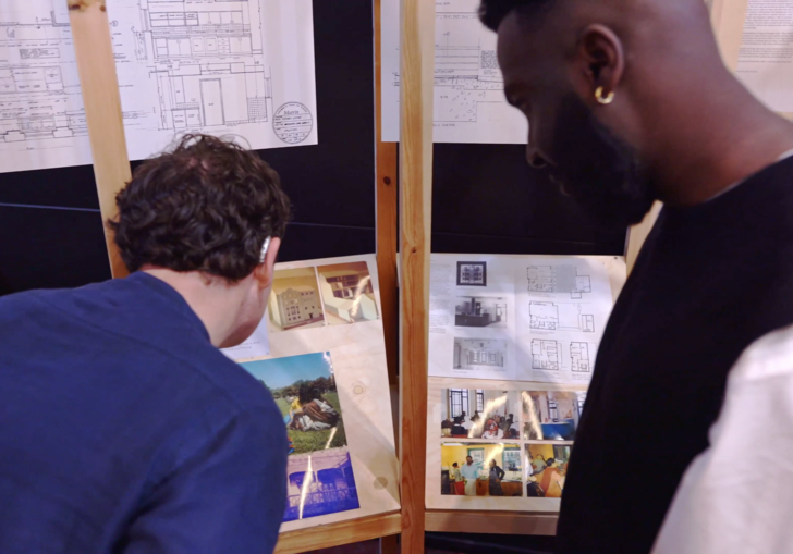 Two people have the back of their heads to the camera, crouching down to look at small printed images of buildings 