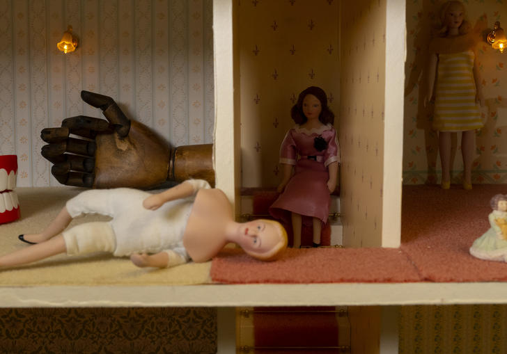 Still image of the interior of a dolls house, some false teeth a wooden hand and two dolls