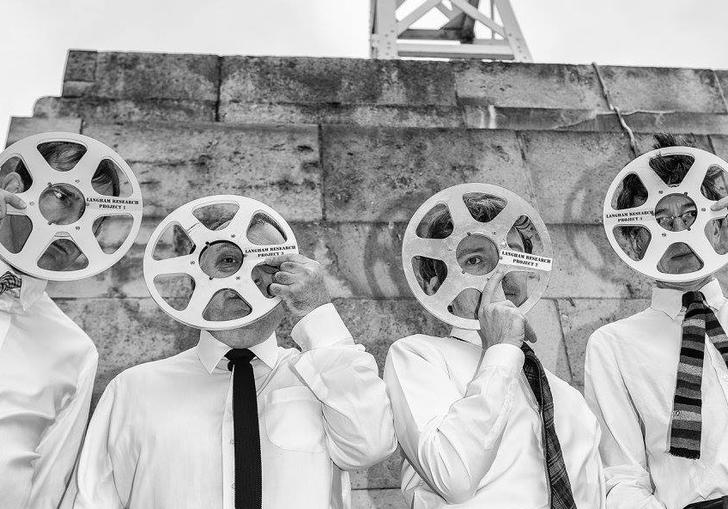 A black and white image of four men dressed in shirts and ties, holding tape reels over their faces