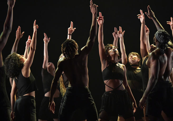 Dancers dressed in black stand together with their hands raised in the hair.