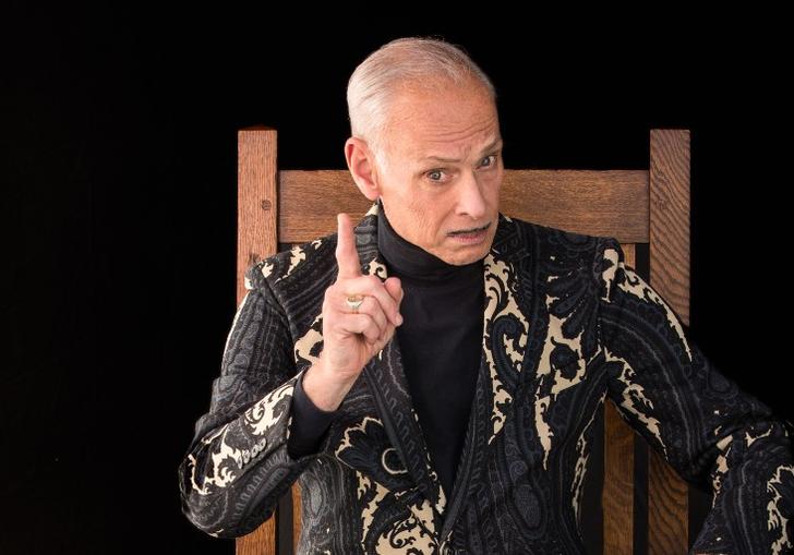 A photo of John Waters sitting on a chair and wagging his finger
