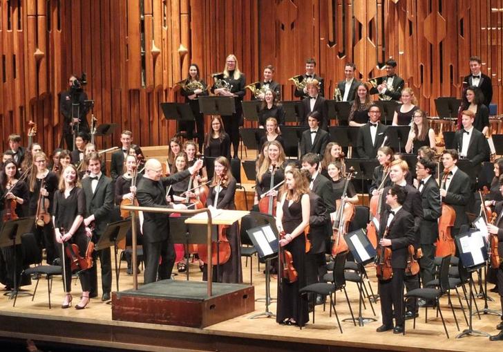 The London School Symphony Orchestra standing on stage in the Barbican Hall holding their instruments