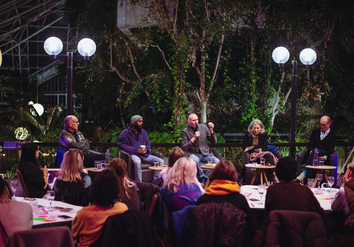 a group of people sit in a row on a stage and an audience of people are facing them with their backs to the camera