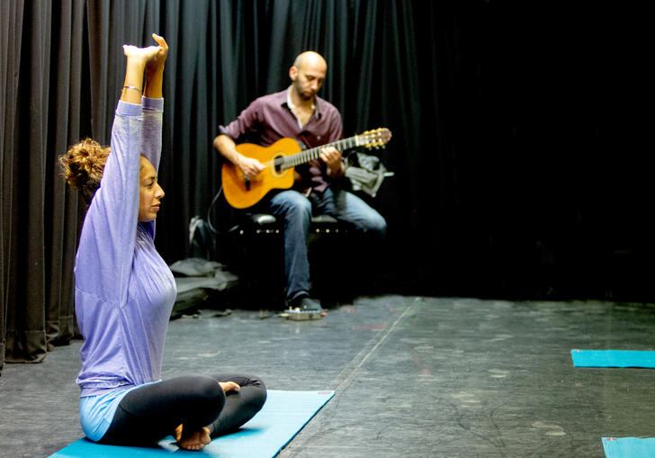 An image from yogabliss workshop: a person is doing yoga, in a cross legged postition, while another person plays guitar to her left 