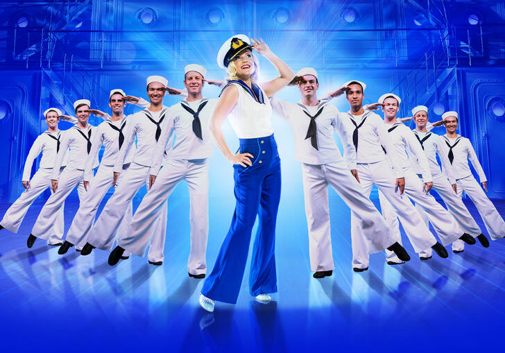 Kerry Ellis dressed in a nautical themed outfit and hat as Reno Sweeney. She stands surrounded by sailors, all saluting. 