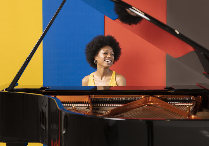 Isata Kanneh-Mason sits at a piano, smiling and looking left. Behind her is a backdrop of yellow, blue, red and black stripes.