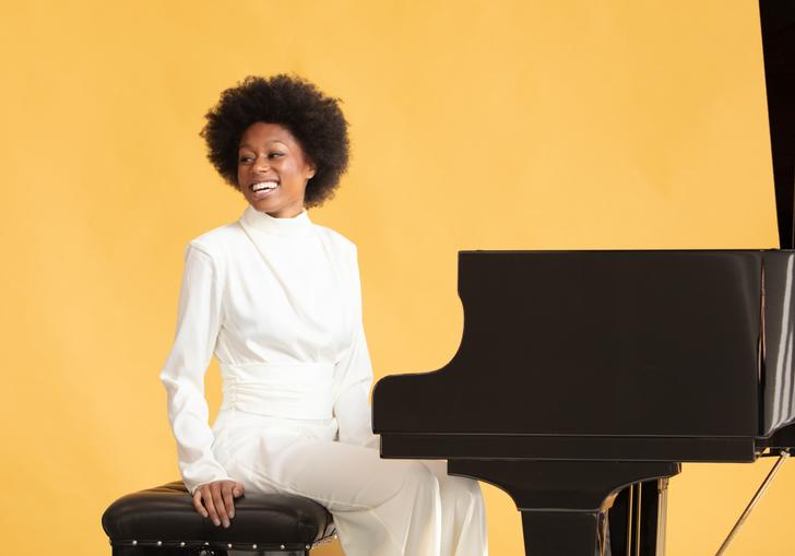 Isata Kanneh-Mason sitting at a grand piano smiling against a pale orange backdrop