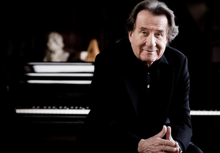 Rudolf Buchbinder sits with his hands touching, with a piano keyboard blurred out behind him.