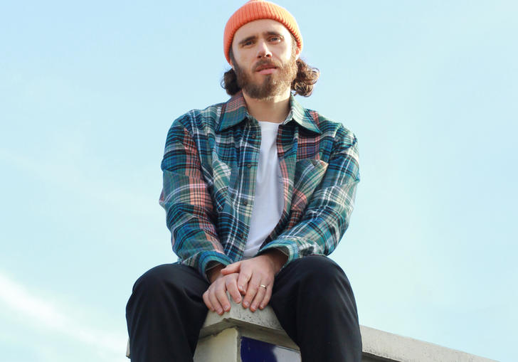 James Vincent McMorrow sitting on the corner of a tiled roof. He is wearing an orange beanie and checked shirt.