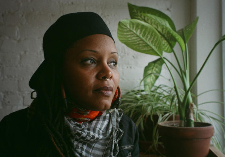 Matana Roberts looking out a window with a plant in front of her. She is wearing a black cap backwards and a white and black dogtooth scarf.