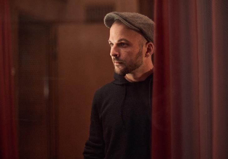 Nils Frahm standing between two red curtains, wearing a flat cap with a black hoodie