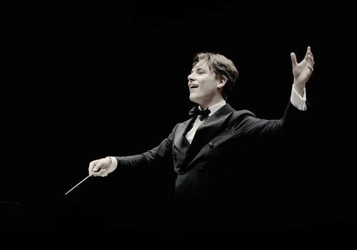 Klaus Makela is conducting in front of a black background, his right hand holding the baton and his left hand above his shoulder