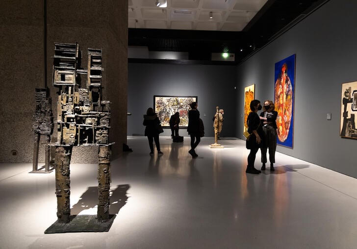 An image of the art gallery with a brown sculpture in the front and people looking at paintings further back from the camera