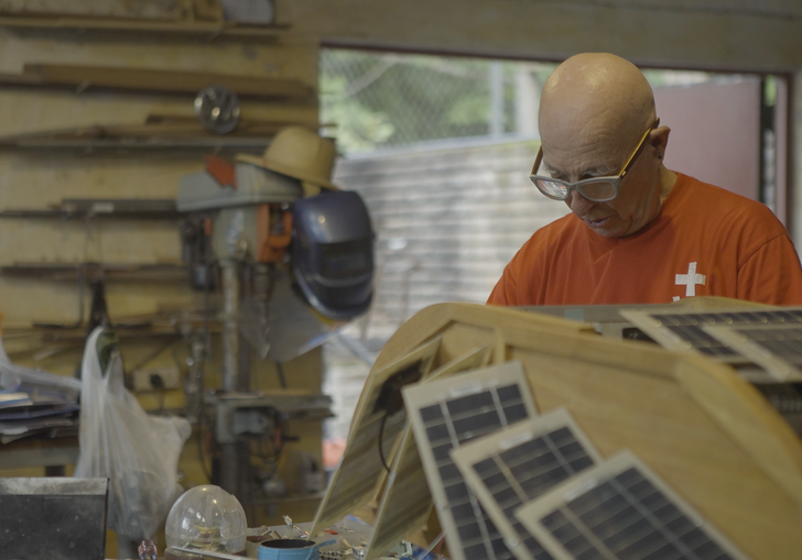 A man in an orange top is in a workshop and a robot with solar panels on the top is placed next to the camera