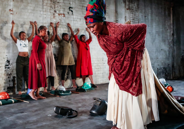A performer in a cream dress and red shawl moves across the stage, behind her a group of 5 performers stand with their arms raised against the wall.