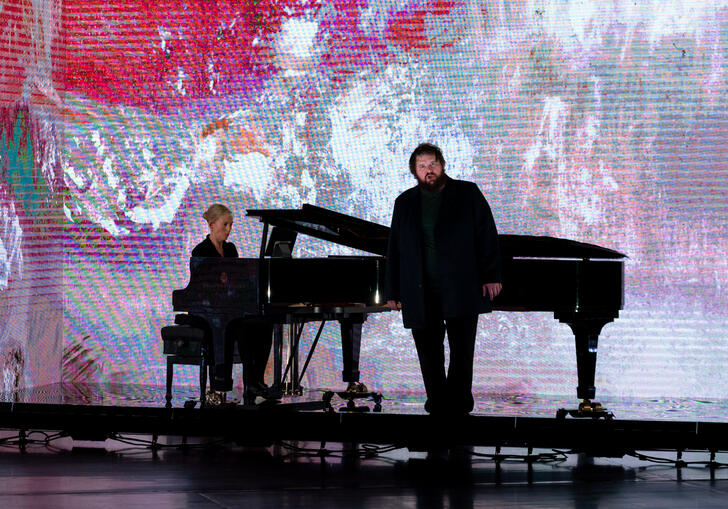 Allan Clayton stands in front of a piano at which Kate Golla sits, with a pink landscape by Fred Williams OBE projected on a screen behind them