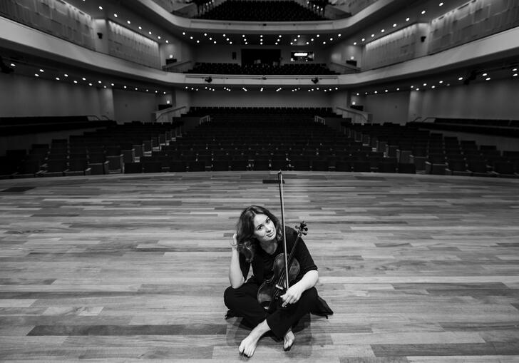 Patricia Kopatchinskaja sitting on a stage holding her violin in her lap