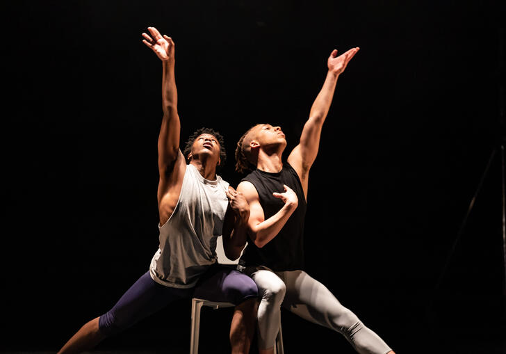 Two Ballet Black performers sit on the same chair with one hand raised towards the sky.