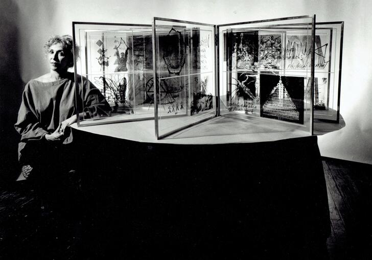 Image of Carolee Schneeman sitting to the left of mutliple screens placed in a circle with photographs
