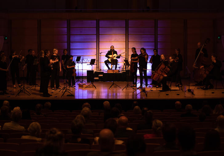 William Barton sits in the middle of the stage on a pedestal playing guitar and didgeridoo. Members of the ACO stand either side of him, in near-darkness