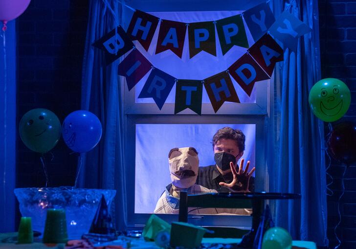 Bill the puppet stares through the window into a room that is dressed for a birthday party.