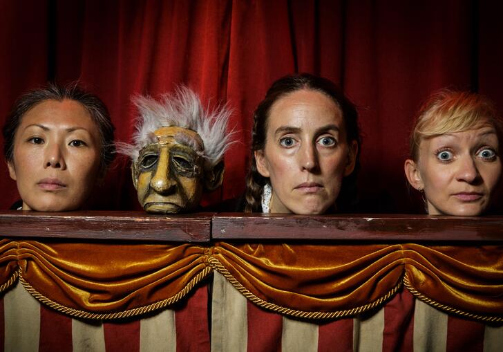 Three performers and one puppet stand in a puppet booth and stare directly at the camera.