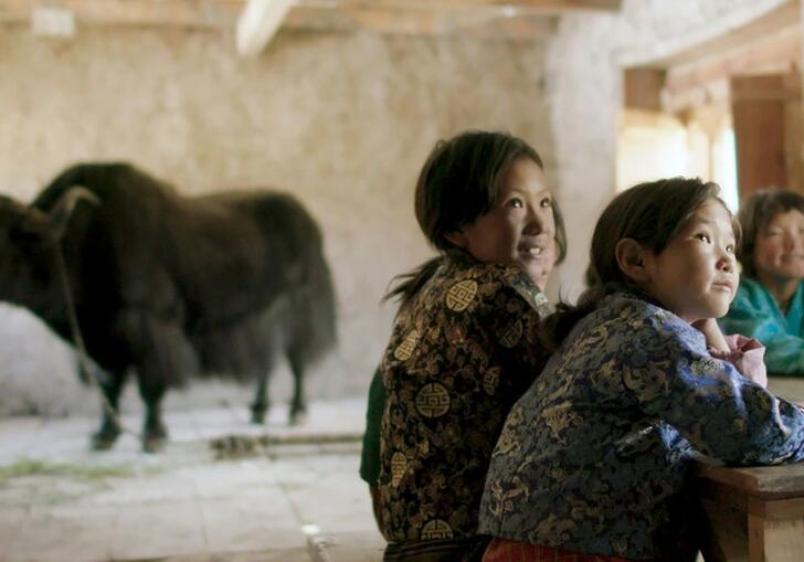 3 children sitting at a table in a classroom looking up. Behind them is a faint black outline of a yak