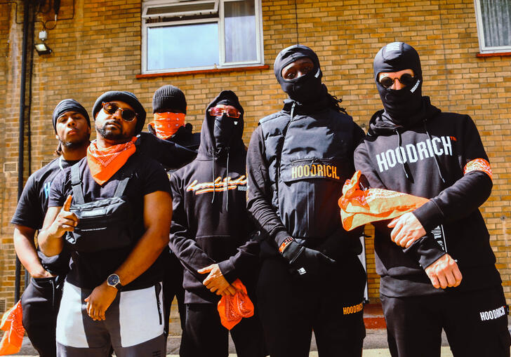 The ensemble dressed in black, some with face coverings, stand in front of some flats and look down the camera.