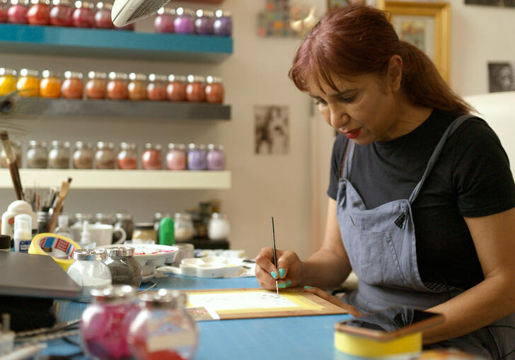 An artist sits at their studio table painting, with an array of multi-coloured paints behind
