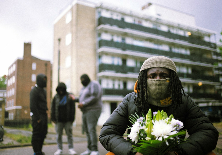 A person whose face is mostly covered with a balaclava holds a bouquet of white flowers. In the backdrop are three others in front of a block of flats.