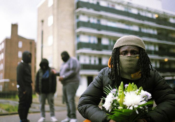 A performer wearing a face covering and holding a bunch of white flowers crouches down in front of a block of flats. Behind them a group of 3 other performers stand talking.