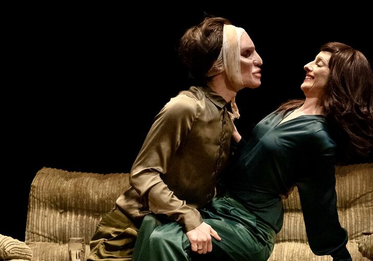 A person wearing a bandage around their head leans over another performer while sitting on a sofa. 