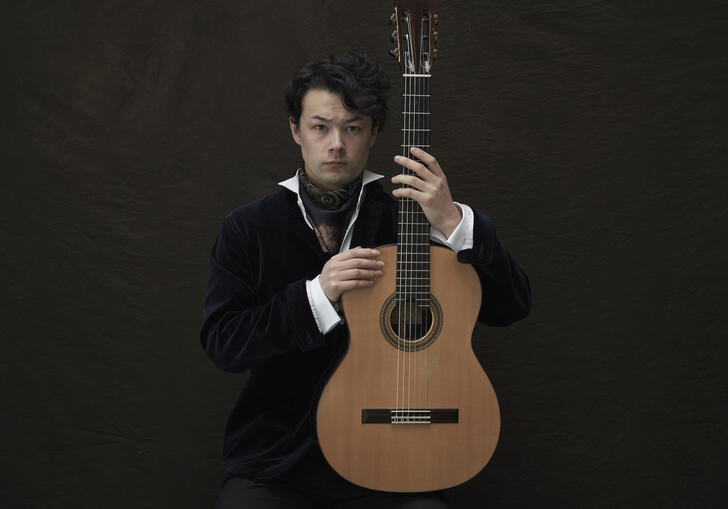 Sean Shibe sitting in front of a dark grey background, holding his guitar upright