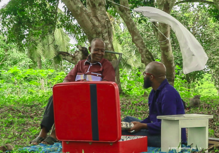 Two black men sit in a forest in front of a red suitcase. Still from Je reste photographe