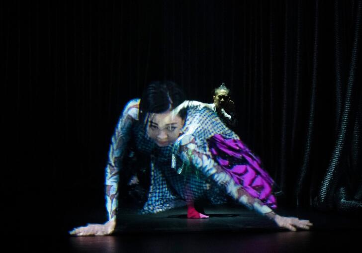 Two performers seem to be merged together while dancing on a stage. One looks like a hologram.