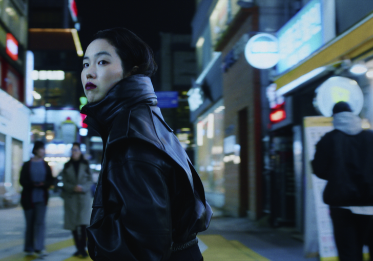A woman in a leather coat standing in a dark shopping street in a city in a still from Return to Seoul