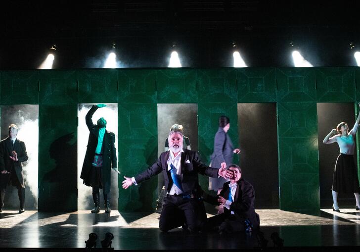 The Cheek By Jowl ensemble perform against a large green structure which has 6 doors cut out of it.