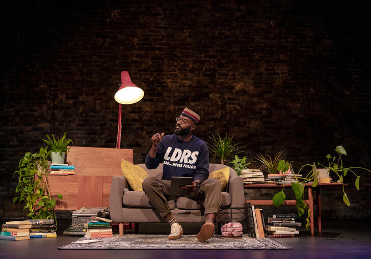 Inua Ellams sits on a stage which is dressed to reflect a living room with a carpet, lamp, table, plants and books.
