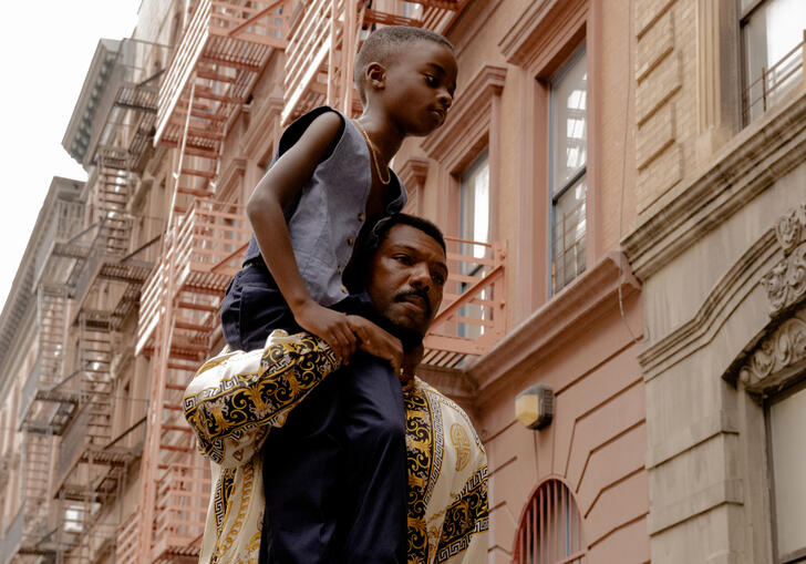A boy sits atop a man's shoulders in a residentual street in a still from A Thousand and One. 