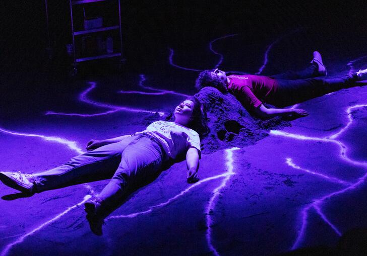 Two people lie on the floor in a dark room. They lie against a mound, which has bright lines coming out of it. They are lit with a purple light.