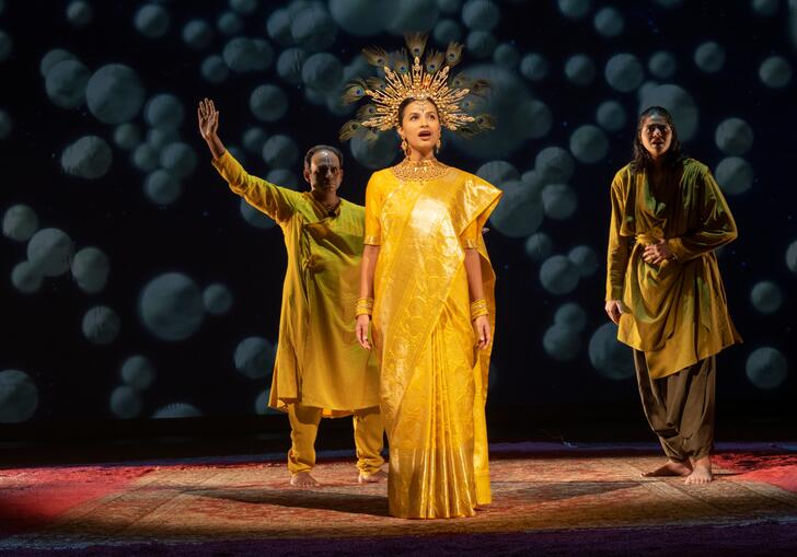 A performer wearing a gold crown sings, while two other performers stand behind.