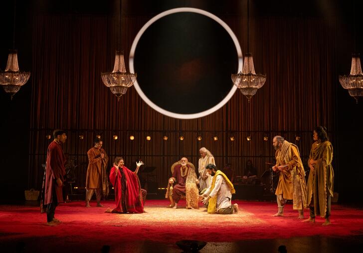 An ensemble of performers gather on stage in a decadent room, a large circle light hangs above them.