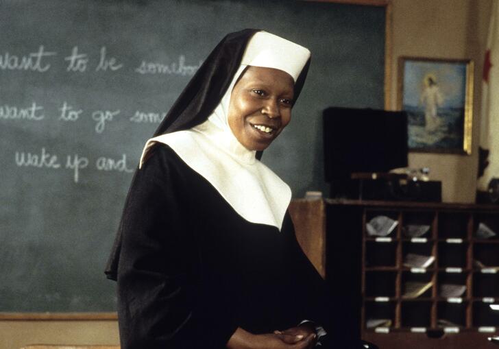 A still from the film Sister Act 2: Whoopi Goldberg is dressed as a nun and sits on a desk in a classroom smiling.