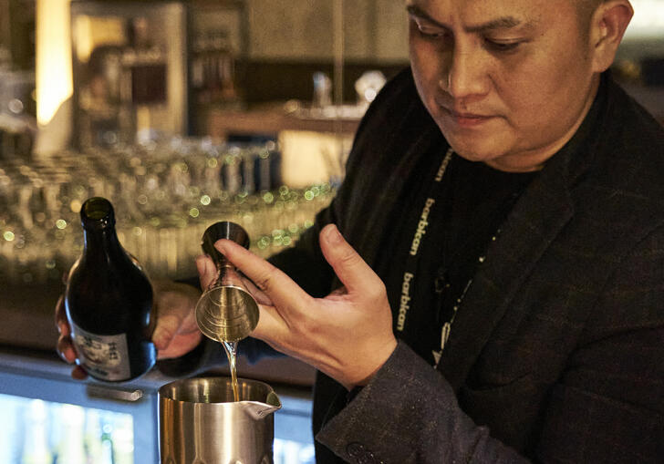 Man pouring a shot of prosecco into a steel jug at a wooden bar.