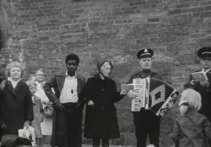 A group of people stand on a stage in the street, reading and playing accordion